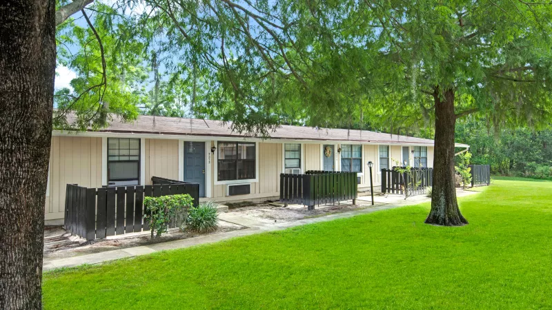Multiple single-story apartments with fenced patios nestled among large trees and green grass.