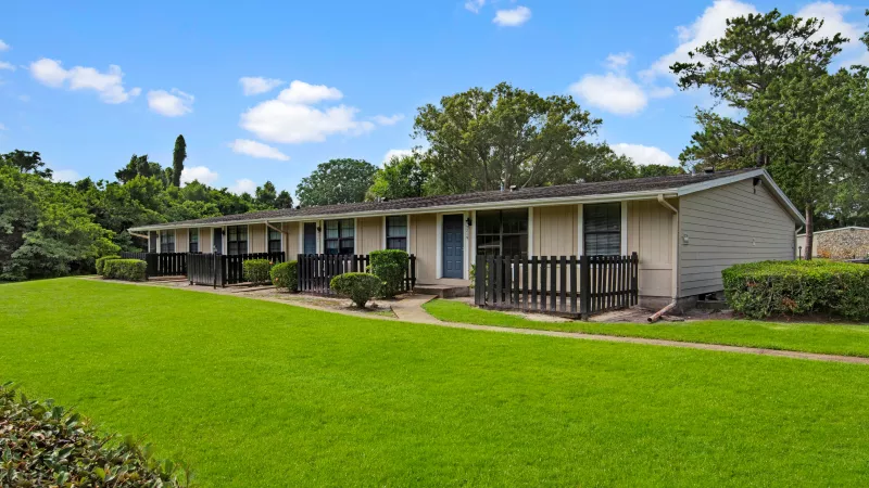 A row of single-story homes with fenced patios and lush green lawns under a bright blue sky.