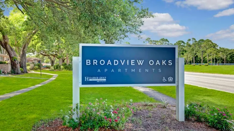 The Broadview Oaks Apartments sign set against a lush green lawn and tree-lined walkway, with a clear blue sky in the background. This image highlights the welcoming entrance and serene environment of the Broadview Oaks community.