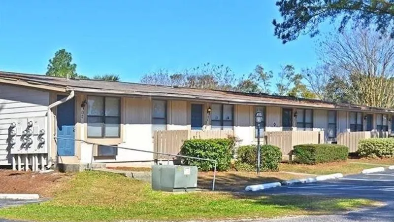 A well-maintained row of single-story apartment units with fenced patios, landscaped greenery, and a nearby parking area under a clear blue sky. This image highlights the practical and serene living environment at Broadview Oaks Apartments.
