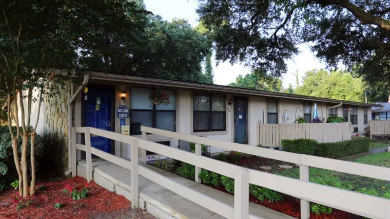 A charming single-story apartment building with a landscaped walkway, wooden railings, and vibrant greenery under the shade of mature trees. This image showcases the welcoming and tranquil atmosphere of Broadview Oaks Apartments.