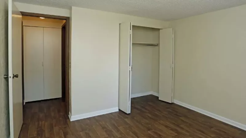 A spacious apartment bedroom with wood-style flooring, neutral-colored walls, and an open closet with bi-fold doors. This image highlights the clean and modern interiors available at Broadview Oaks Apartments.