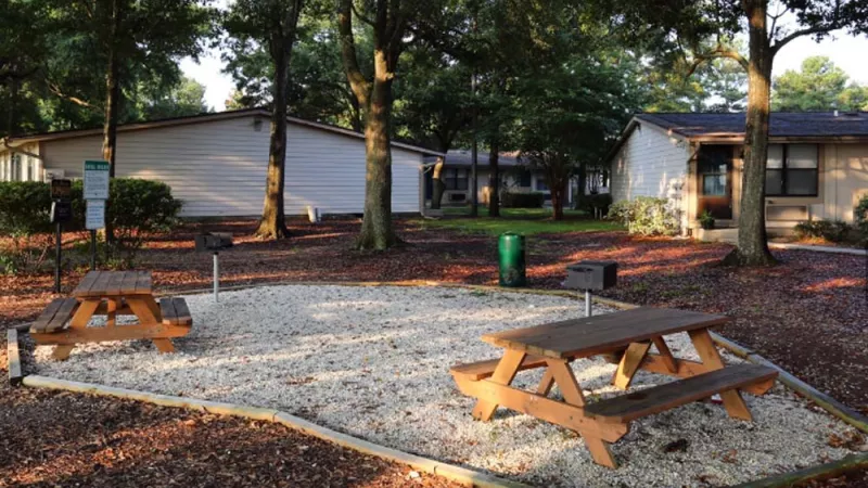 Alt description: A serene outdoor picnic area with wooden tables, gravel ground cover, and barbecue grills, surrounded by tall trees and well-kept apartment buildings. This image highlights the relaxing community amenities at Broadview Oaks Apartments.