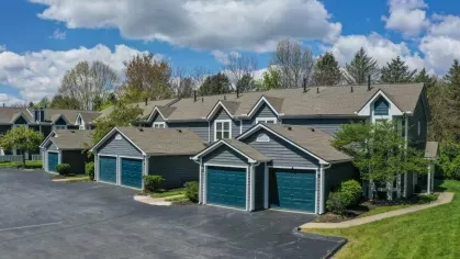 Exterior view of a modern apartment complex with attached garages, set against a bright blue sky with scattered clouds. The well-maintained landscaping and spacious parking area enhance the community's curb appeal.