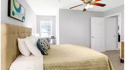 A bright and spacious bedroom with soft carpeting, neutral wall colors, and a ceiling fan, leading to a walk-in closet.