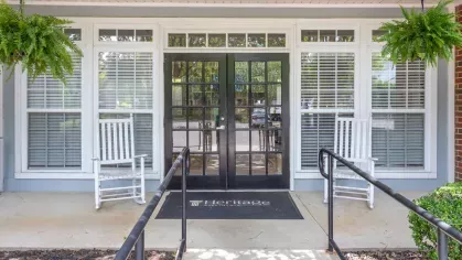 Entrance with white rocking chairs and glass-paneled doors surrounded by vibrant greenery at Heritage Apartments.