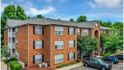 Side angle of a three-story red brick apartment building at Heritage Apartments, with adjacent parking spaces and well-maintained landscaping.
