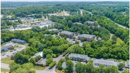 Wide aerial view of the Heritage Apartments community and surrounding areas, including nearby retail and wooded spaces.