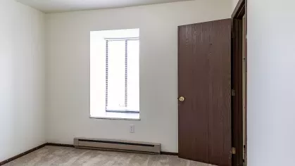 A bright and spacious bedroom with natural light pouring through a large window, complemented by clean beige walls and plush carpeting, ideal for comfort and relaxation.