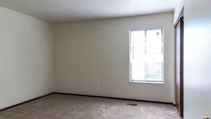 A bright and spacious bedroom with natural light pouring through a large window, complemented by clean beige walls and plush carpeting, ideal for comfort and relaxation.