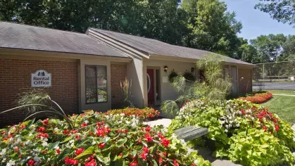 The rental office at North River Place Apartments, surrounded by colorful flower beds and shaded by mature trees