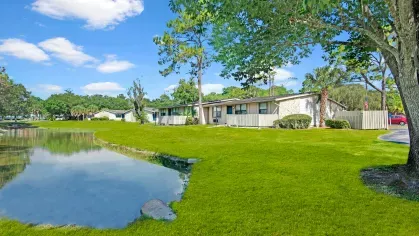 A serene lakeside view of single-story apartments with green lawns and tall trees.