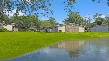 A serene view of single-story apartment buildings surrounded by lush greenery and a reflecting pond.