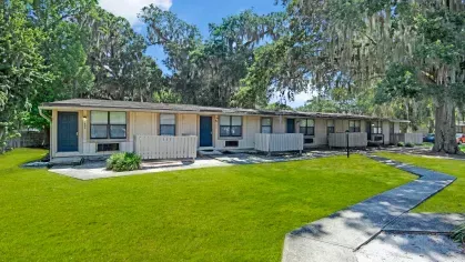 A cluster of single-story apartments set amidst vibrant green lawns and shaded by tall trees.