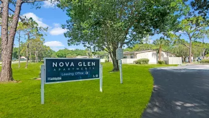 The Nova Glen Apartments sign surrounded by lush landscaping and shaded by tall trees.