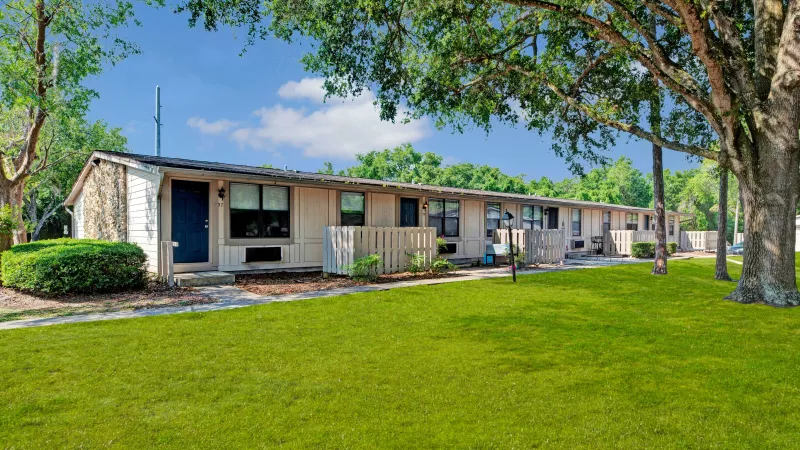 A row of light beige single-story apartments surrounded by vibrant green grass and trees on a sunny day.