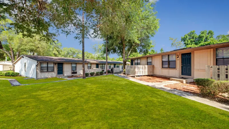 Multiple beige apartments with blue doors, surrounded by well-maintained greenery and a spacious yard.