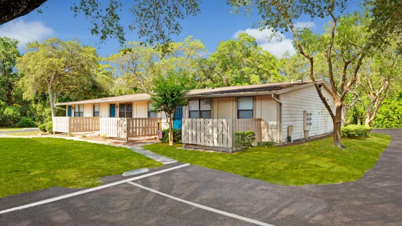Beige apartments with picket fences, set near a parking lot and shaded by trees on a clear day.