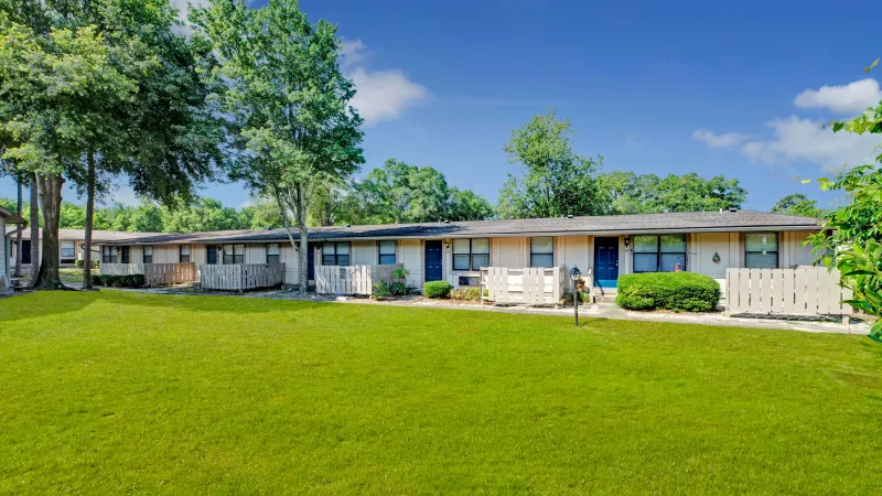 A wide-angle view of beige apartments with blue doors, neatly aligned amidst greenery and pathways.