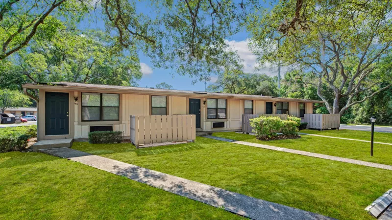 A small residential unit with a fenced patio, surrounded by trees and grass under a clear blue sky.