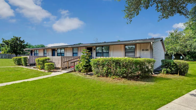 A single-story residential building with beige siding, blue doors, and neatly trimmed bushes in the foreground.