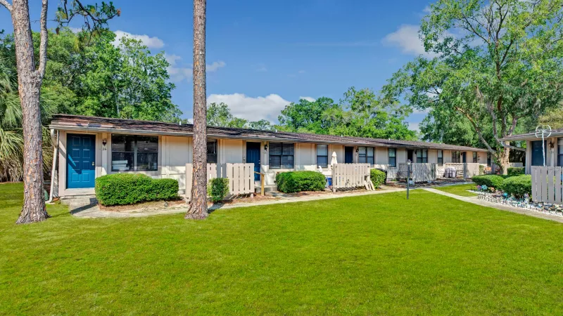 A row of single-story residential units with white picket fences and green lawns, surrounded by tall pine trees.