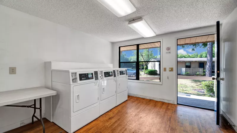 A laundry room with several white machines, a folding table, and large windows overlooking the outdoor area.
