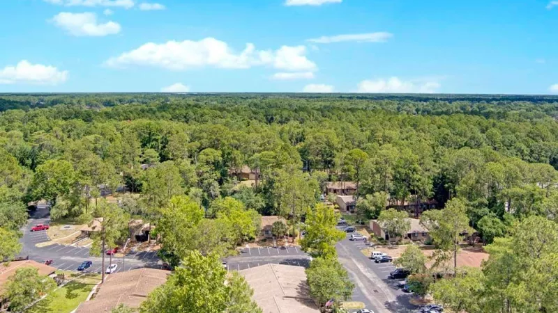 An aerial view of a residential community surrounded by dense, lush green forests under a bright blue sky.