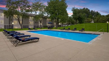 Outdoor swimming pool surrounded by lounge chairs, with a sunset sky and a grassy area in the background.