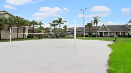 A sandy volleyball court surrounded by green grass, palm trees, and residential homes under a blue sky.