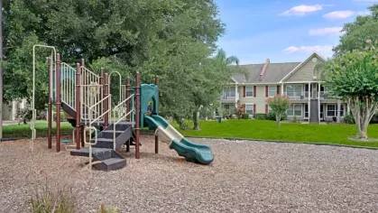 A children's playground featuring slides and climbing structures, with trees and residential buildings in the background.