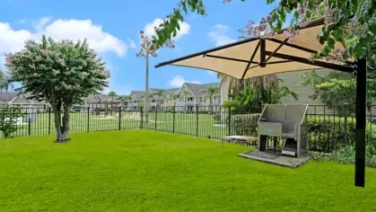 A fenced dog park with shaded seating, a barbecue station, and a well-maintained green lawn.