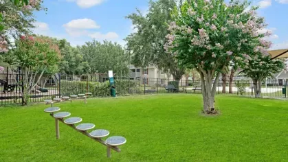 A grassy outdoor space with a balance beam play structure, blooming trees, and a playground in the background.
