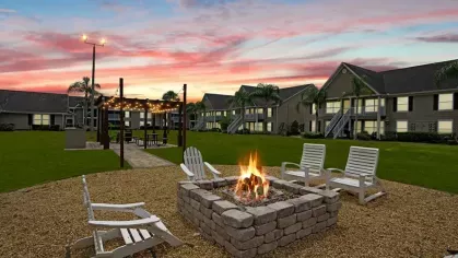 A cozy fire pit surrounded by Adirondack chairs, with string lights and residential buildings under a colorful sunset.