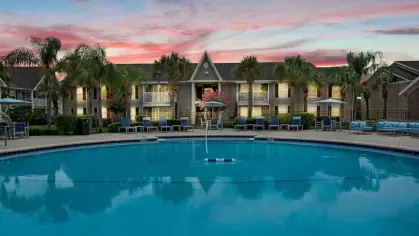 A sparkling pool area surrounded by palm trees, lounge chairs, and residential buildings at sunset.