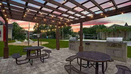 A covered outdoor picnic area with string lights, barbecue grills, and seating, set against a colorful sunset sky.