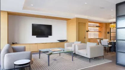 A modern library with wooden shelving filled with colorful books, two circular chairs, and a small round coffee table in a well-lit space with large windows.