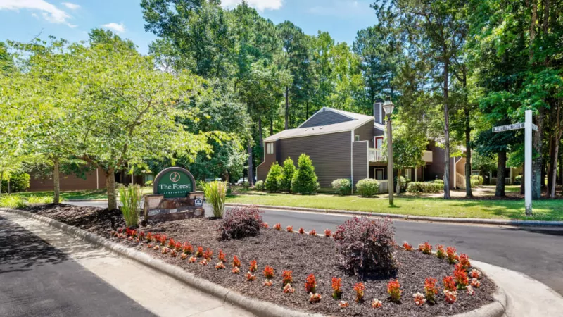The entrance to "The Forest Apartments," featuring landscaped greenery, vibrant flowers, and modern buildings surrounded by tall trees.