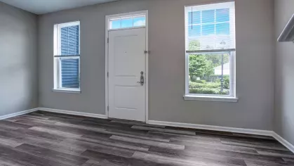 An open living area with hardwood flooring, light gray walls, and a large front door with side windows offering views of the outdoor greenery.