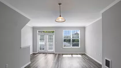 An open living area with hardwood flooring, light gray walls, and a large front door with side windows offering views of the outdoor greenery.