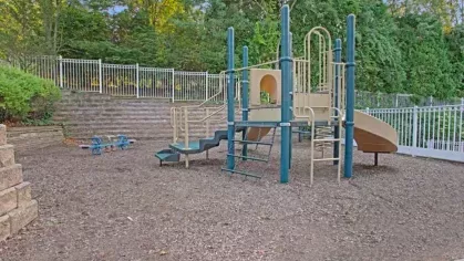 A playground area with slides, climbing structures, and seating, surrounded by a landscaped and fenced setting.