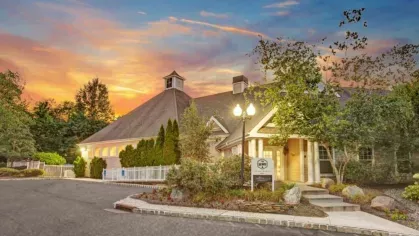 A clubhouse building with warm lighting at dusk, featuring a landscaped entrance and a sunset backdrop.