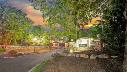 A gated community entrance with a sign, surrounded by lush trees and landscaping at sunset.
