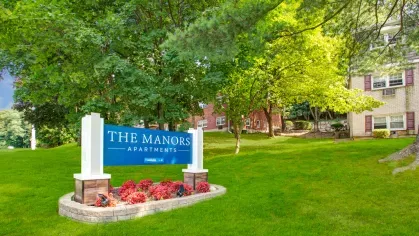 The Manors Apartments sign displayed on a beautifully landscaped area with vibrant flowers and mature trees.