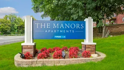 Prominent blue and white signage for The Manors Apartments set against a vibrant green lawn and landscaped flower beds, highlighting the community’s attention to detail and curb appeal.