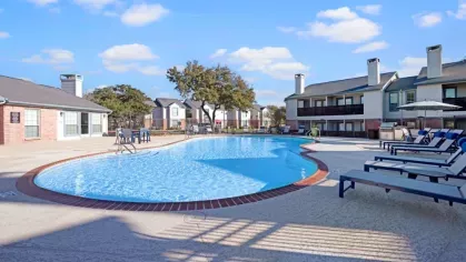 A sparkling swimming pool surrounded by lounge chairs, shaded seating areas, and beautiful apartment buildings at The Maxwell Apartments in San Antonio, TX.