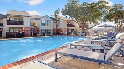 A sparkling swimming pool surrounded by lounge chairs, shaded seating areas, and beautiful apartment buildings at The Maxwell Apartments in San Antonio, TX.