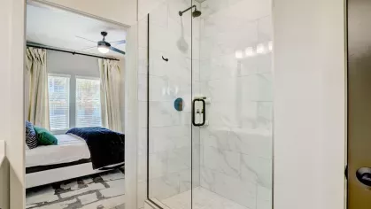 A close-up of a sleek glass-enclosed shower with marble tile, adjacent to a bright and airy bedroom view.