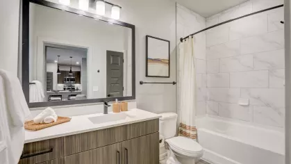 A spacious bathroom with a modern vanity, large mirror, and a tiled shower-bath combination, reflecting the open-plan kitchen in the mirror.