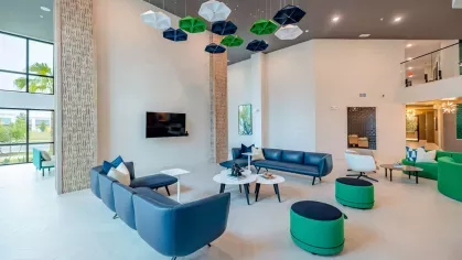Image 5: Inviting community lounge with bold green and blue seating, modern decor, and ample space for gatherings at Tropia Wellen Park Apartments.